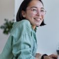 Smiling woman in glasses and green shirt working on a laptop at a desk in a bright, modern office or co-working space.