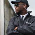 A male security guard leaning on a glass door with his arms crossed. He's wearing a hat, sunglasses, and an earpiece.