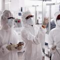 Three adults in white hazmat suits, gloves, and masks, standing inside a modular room with glass walls.