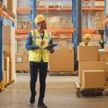 A warehouse worker in a safety vest and hard hat holding a tablet and surveying rows of cardboard boxes.