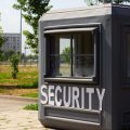 A dark gray security booth sits unattended on a paved area next to a parking lot. A hotel is in the distance.