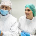 Two laboratory workers in white protective suits and blue gloves looking at equipment inside a large room.