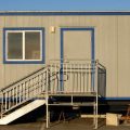 A gray and blue portable office with metal stairs sits in a warehouse or construction site parking lot.