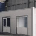 A white portable office featuring doors and windows is in the corner of an industrial facility with metal walls.