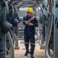 An person wearing blue coveralls and a hard hat walks through a room with HVAC equipment. He is holding a tablet.