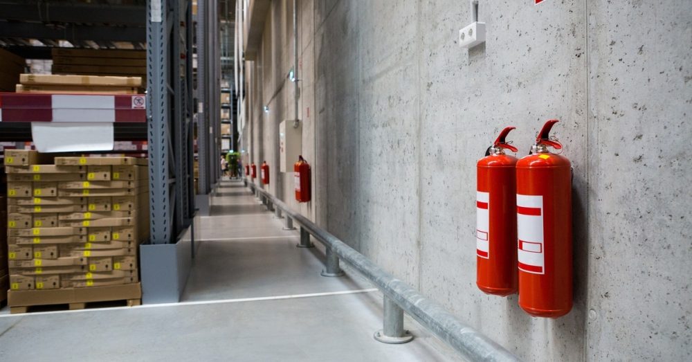 Several fire extinguishers hang on a concrete wall extending across a warehouse. Aisles with products can be seen.