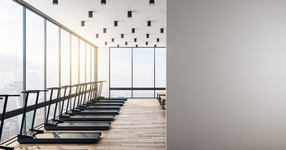 A gym with open windows has a blank wall that separates the space. One side of the room has a row of treadmills.