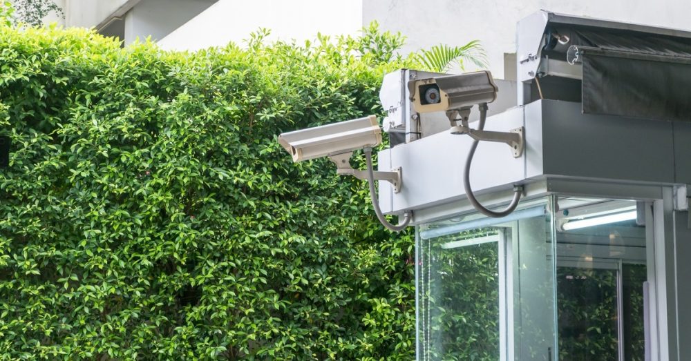 Two security cameras are installed on the roof of a guard booth outside a large building. A hedge separates the area.