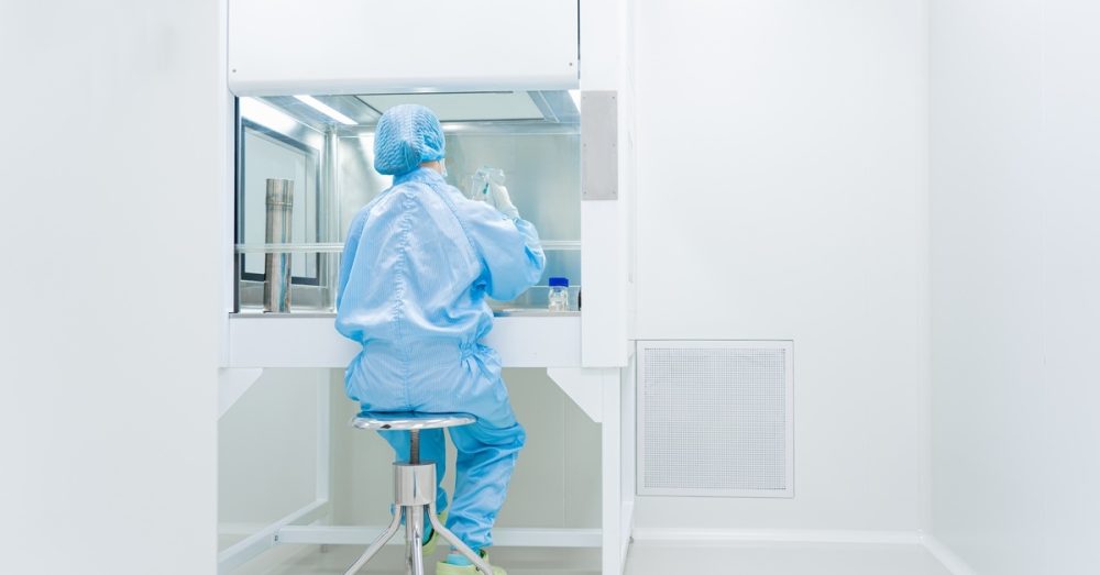 A scientist sits on a stool in a brightly lit cleanroom as they test samples in a laboratory workspace.