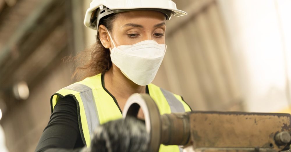 A woman in a factory operates a metal machine by spinning a lever. She wears a hard hat and a face covering.