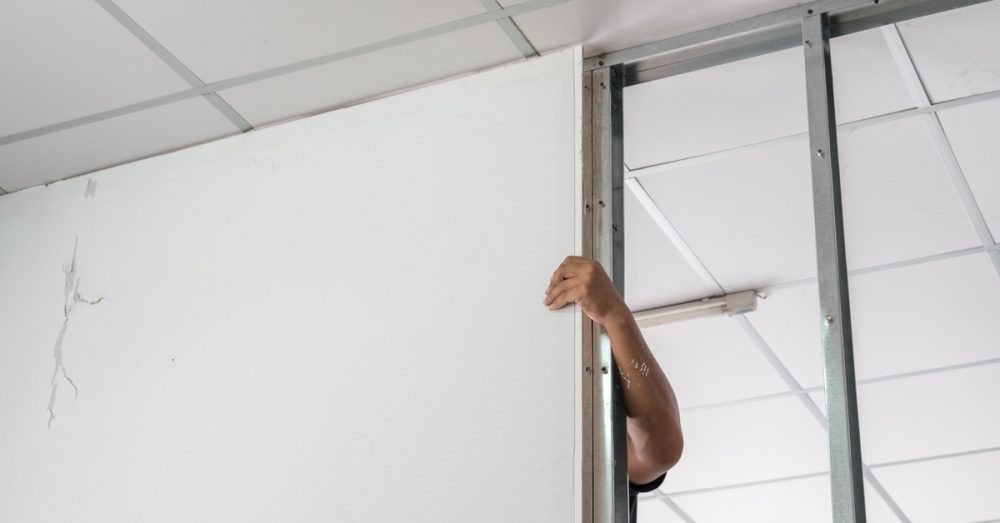Worker's arm holding a white partition wall inside a building with metal framing and a tiled ceiling.