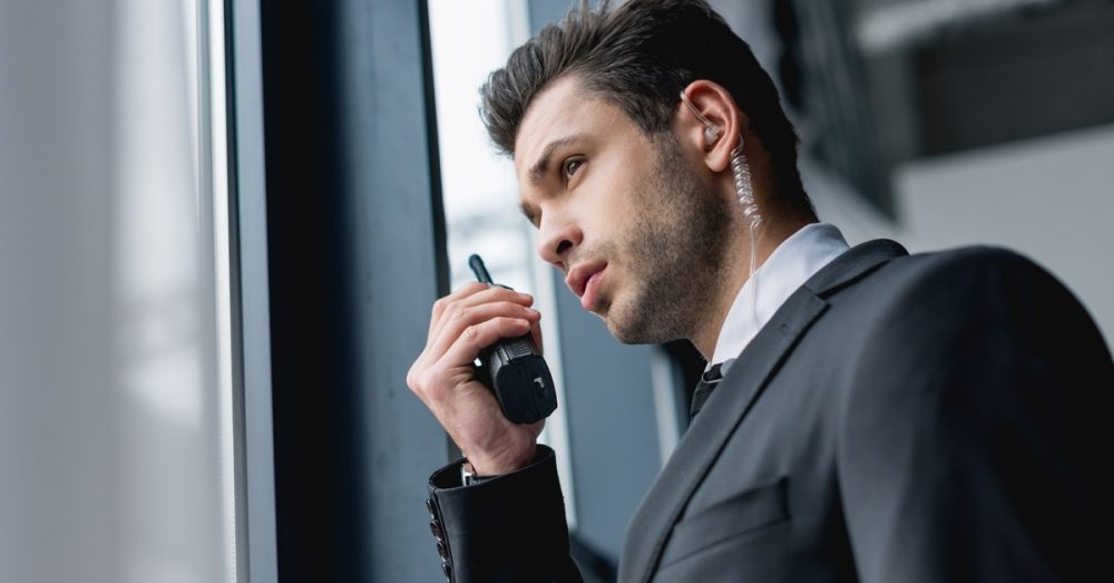 A young man holding a two-way radio and looking out a window. He's wearing a suit and has a clear earpiece.