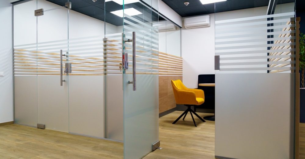 A modern office with frosted glass partitions and wood flooring. A yellow chair is visible through an open partition door.
