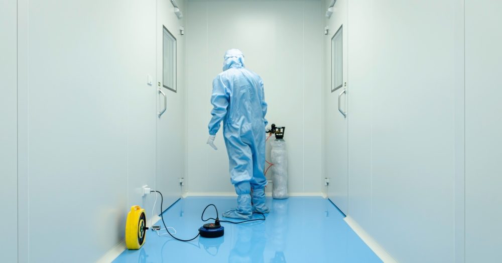 A technician in a blue hazmat suit and white gloves cleaning the floor between two clean room doors.