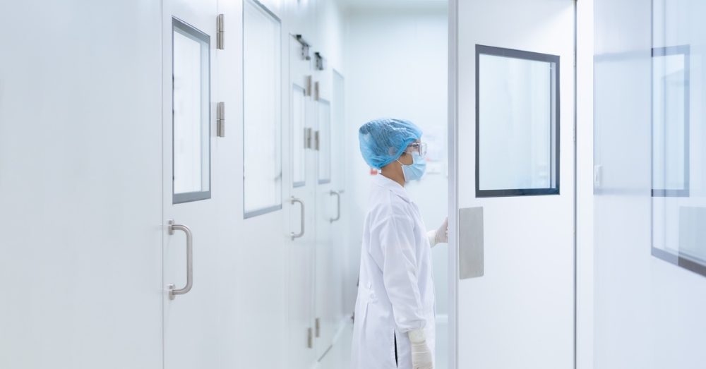 A woman in a lab coat and gloves opening the door to a clean room. The walls, floors, and ceiling are all white.