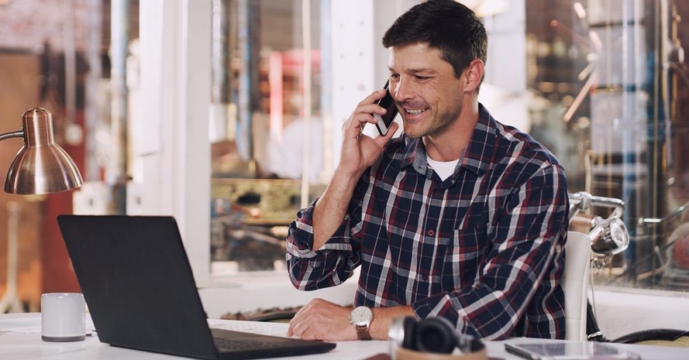A middle-aged man holding a cell phone and looking at a laptop on a desk inside a glass-walled warehouse office.