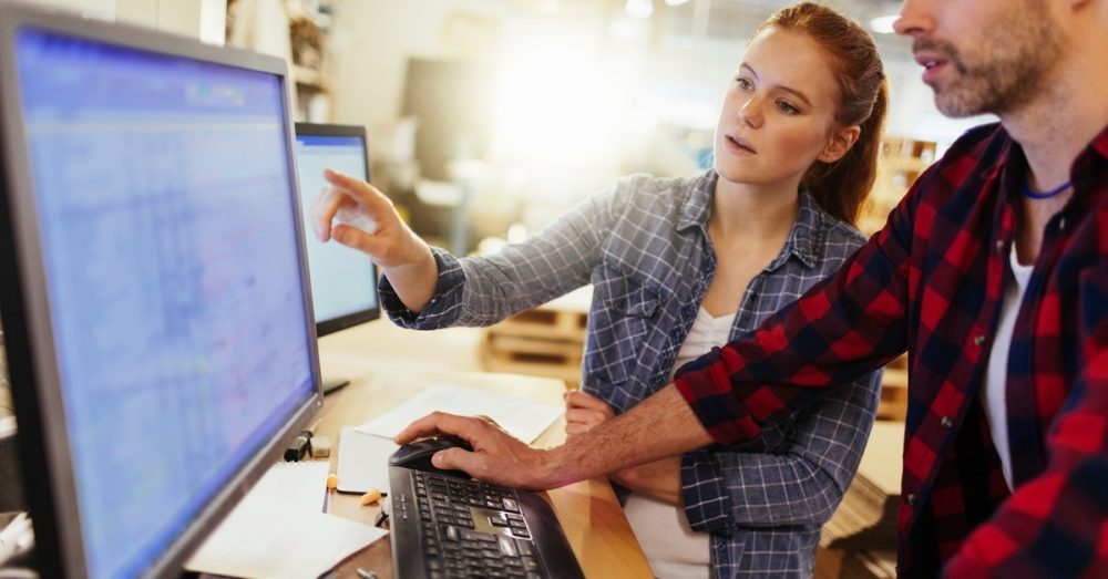 Two young adults standing and working on a computer inside a well-lit warehouse full of wooden pallets.