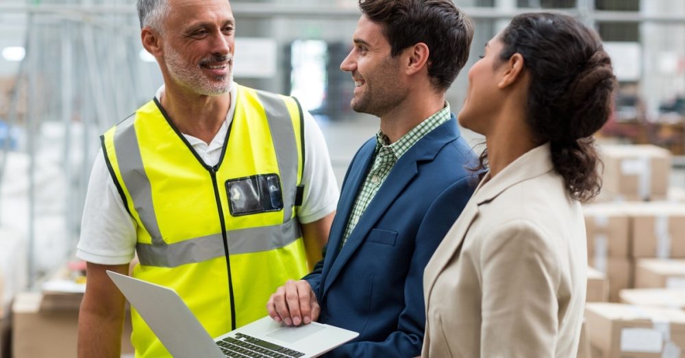 Two adults in blazers standing inside a warehouse and turning to speak to a senior man in a safety vest.