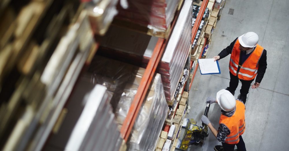 A male warehouse worker positions a pallet jack under a large shelf while another holds a blue clipboard.