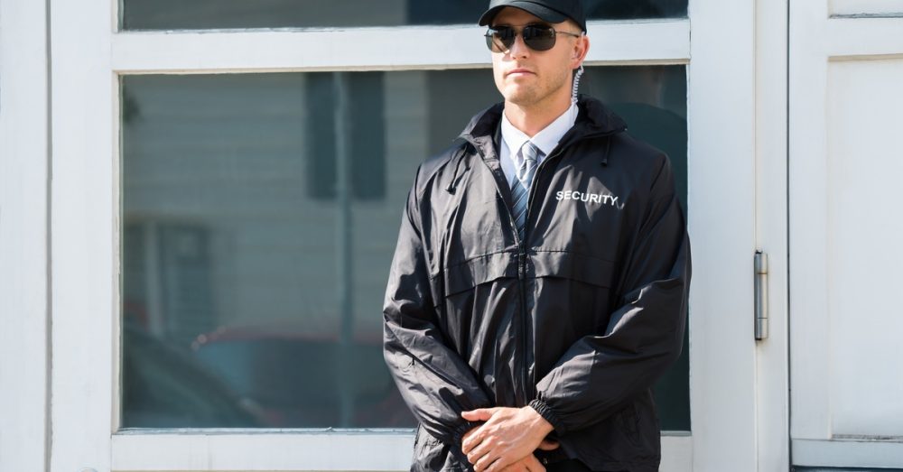 A man in a black security uniform stands with his hands in front of him near the entrance to a building.