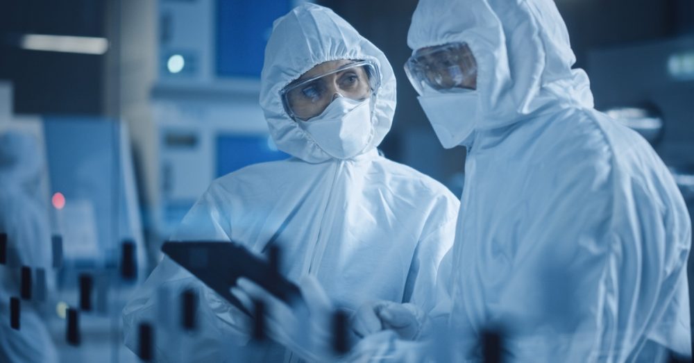 Two scientists in full protective hazmat suits and goggles examining data on a tablet in a sterile laboratory.