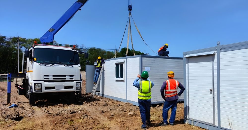 A truck with a forklift connected to a portable trailer office with a window cutout. Several workers huddle around the unit.
