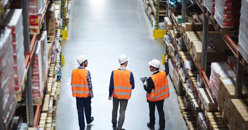 A group of warehouse workers wearing white hard hats and orange vests talk as they walk between aisles of a warehouse.