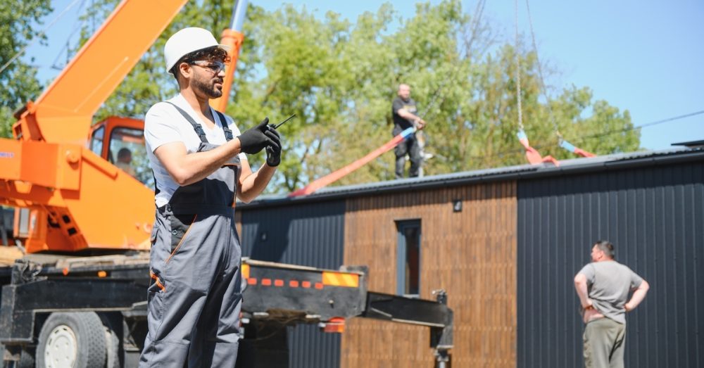 A man wearing a hard hat and overalls stands in front of a crane dropping a modular building onto a construction site.