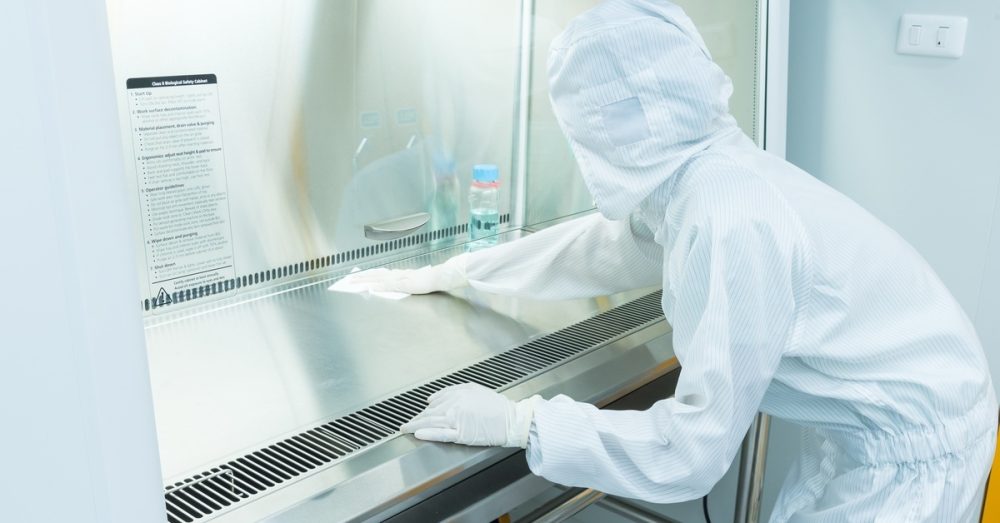 A scientist in full protective body suit gear wiping down a stainless steel work surface in a clean room.