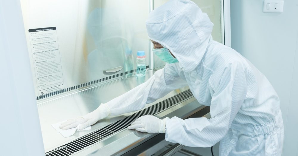 A person in a full-body protective suit and mask leans over a safety cabinet, wiping it down with a tissue and cleaning solution.