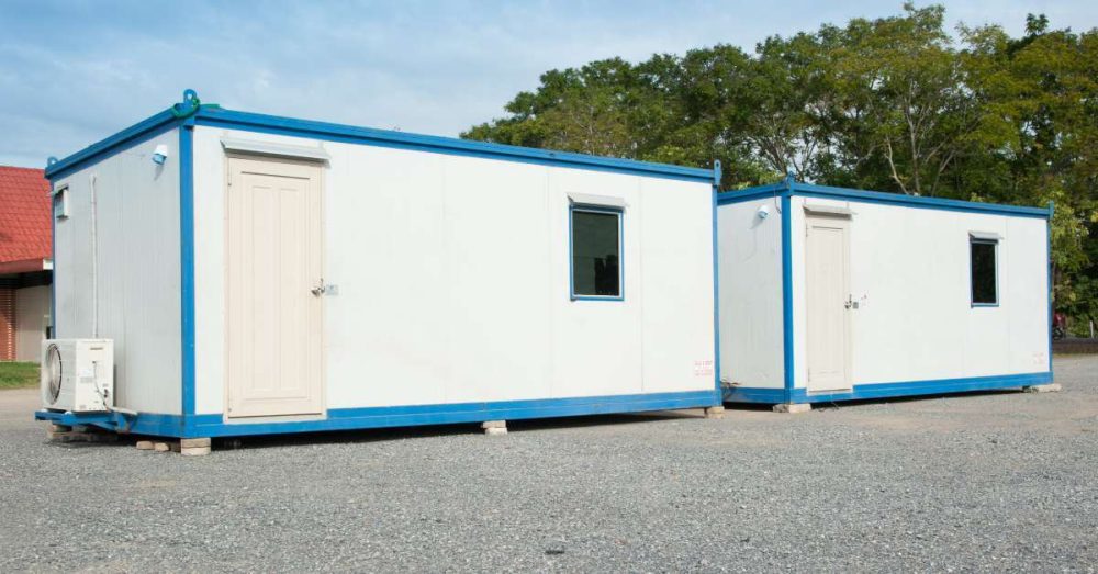 Two white and blue portable offices sit side by side in a parking lot. They are each resting on top of bricks.