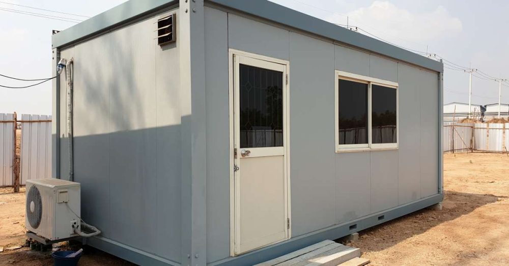 A gray mobile office building on an unfinished lot. There are shoes sitting outside of the front door.