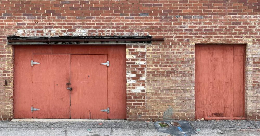 A large brick wall with two red wooden doors and three windows. The warehouse exterior seems worn and faded.