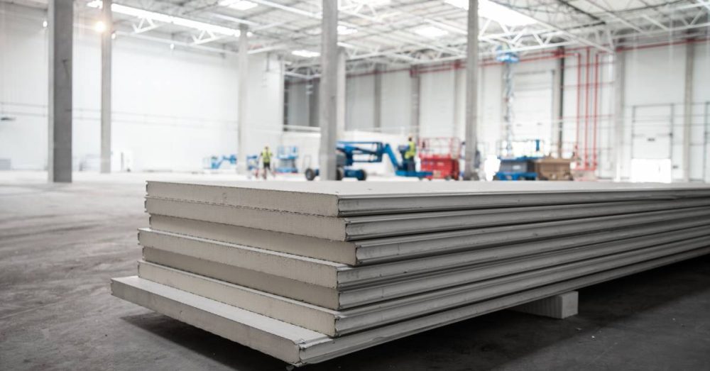 A large warehouse nears completion as two workers inspect it. Several materials are stacked at the front of the image.