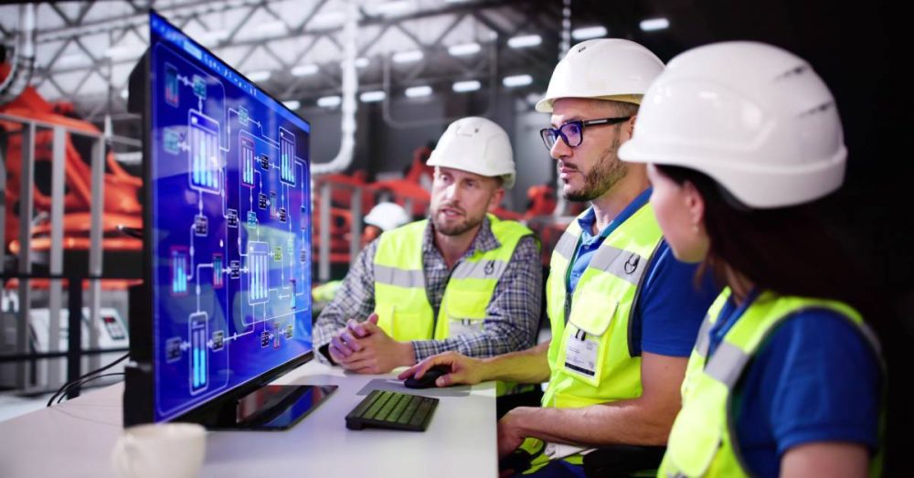 Three workers gather at a computer to analyze the systems of their industrial plant. There are schematics on the screen.
