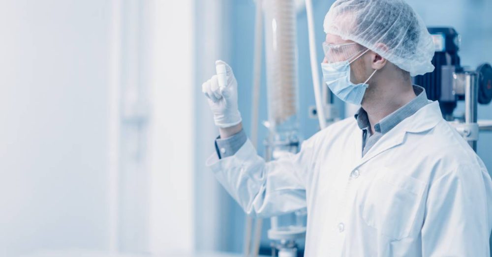 In a laboratory, a scientist wearing protective glasses, a hairnet, and gloves holds a small product at eye level.
