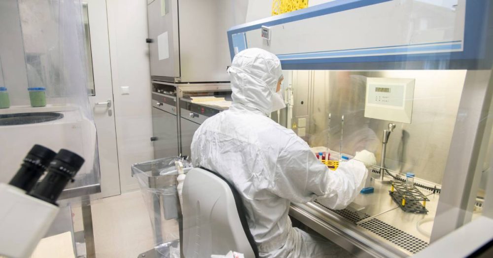 A scientist sitting in a chair while wearing a body suit. The laboratory has several pieces of equipment on the table.