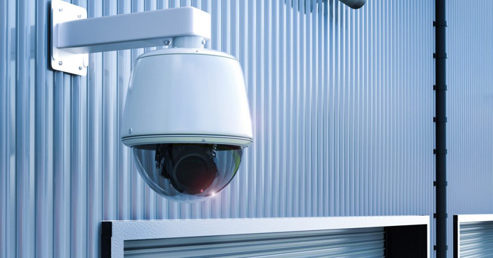 A domed security camera and spotlights on a gray-blue exterior steel wall above a closed loading bay door.