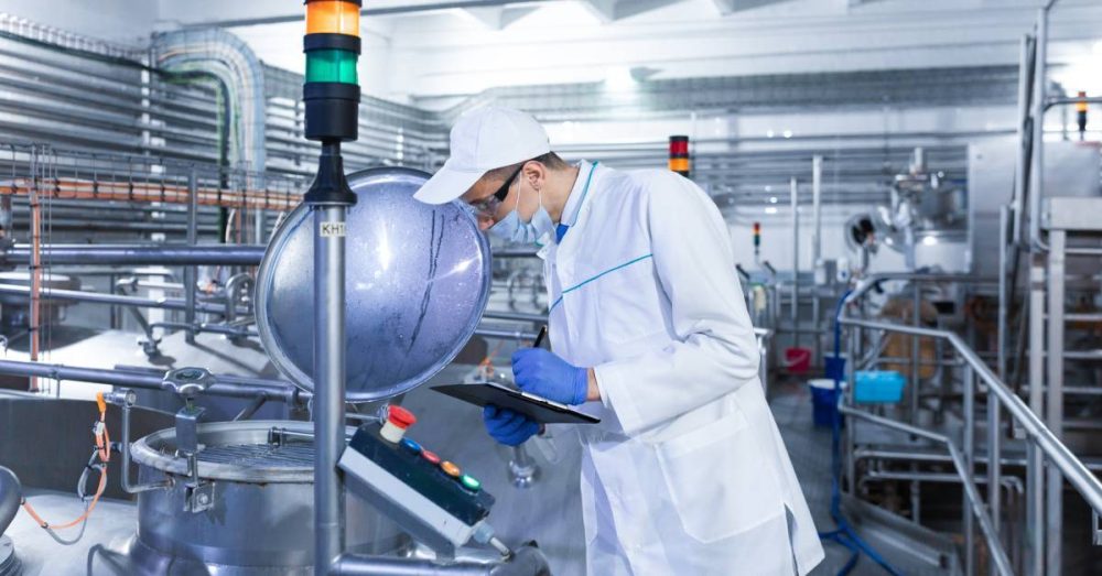 A man wearing a white robe and mask takes notes on a clipboard. He is standing in front of machinery.