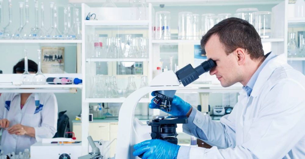 A scientist wearing blue gloves peers into a microscope. Another scientist stands in the background of the lab.