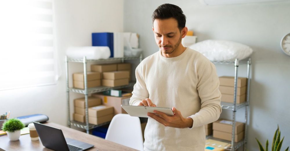 A man standing next to a desk and using a silver tablet. There are boxes and indoor plants behind him.