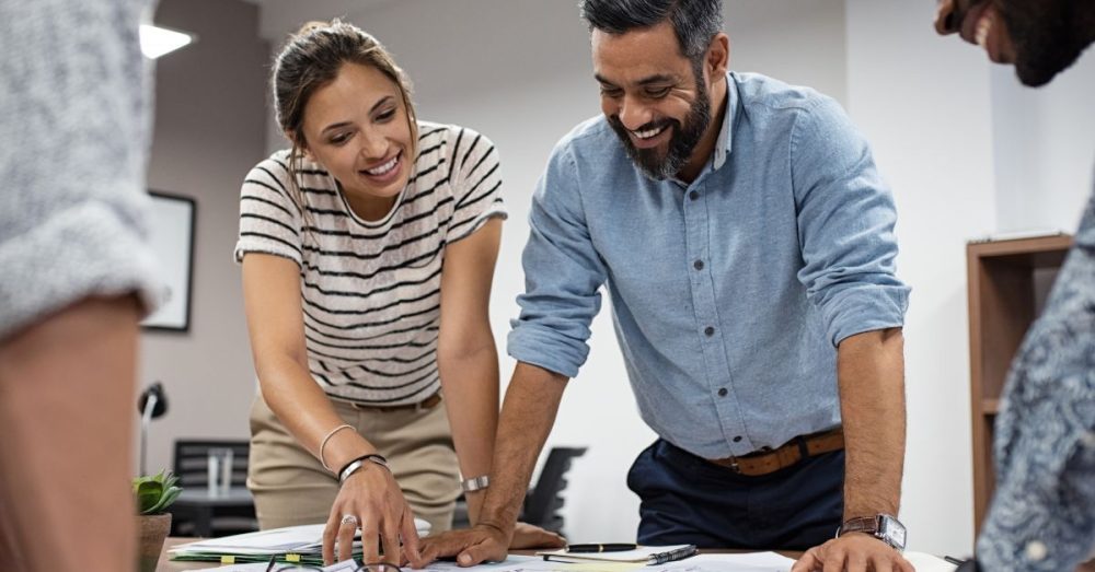 A group of smiling people gathered around a large table. They're looking at multiple building blueprints.