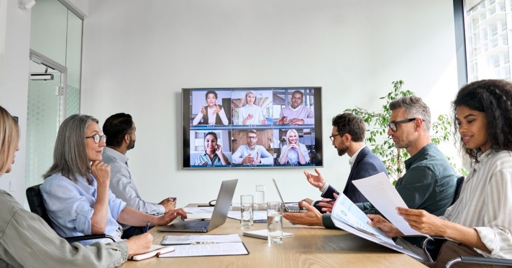 A group of six people gathered around a desk in an office meeting room, with six more people on a call on a screen in the room.